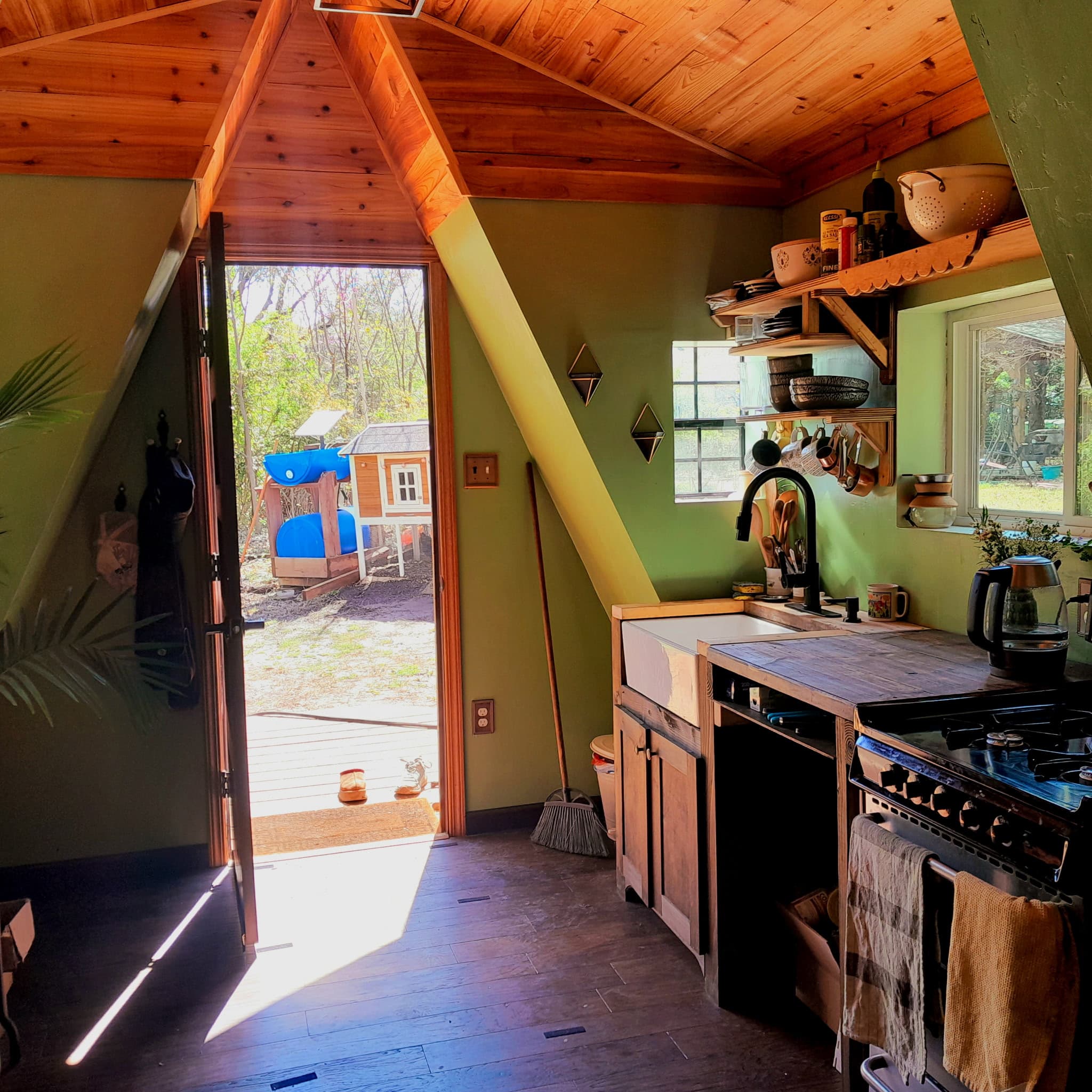 Inside the tiny house — wood-panel A-frame ceiling, sage walls, kitchen with farmhouse sink and gas range